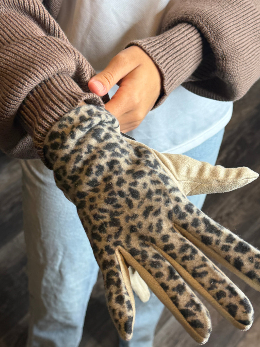 Person wearing a leopard print glove with a brown sweater on a wooden floor.