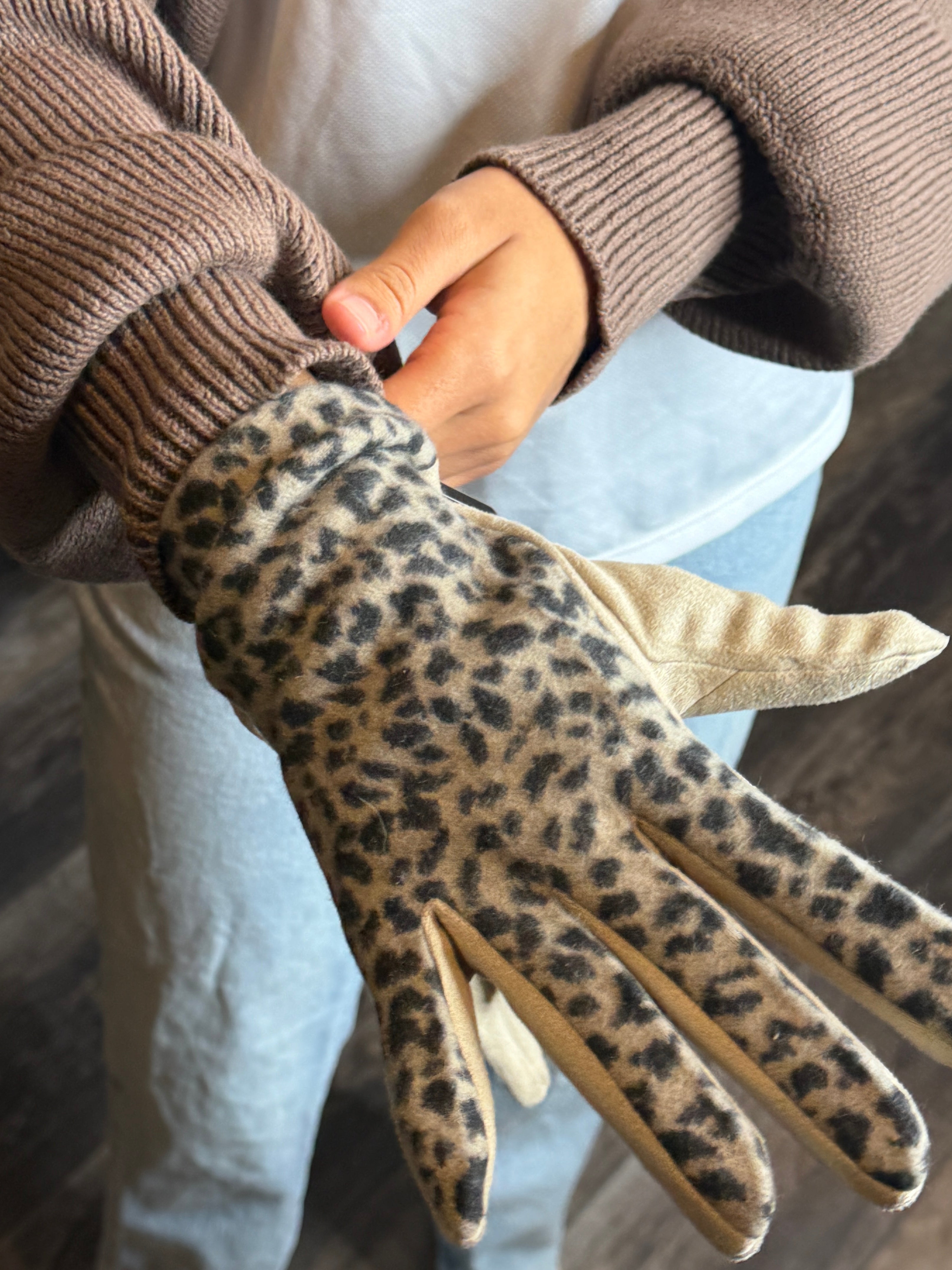 Person wearing a leopard print glove with a brown sweater on a wooden floor.